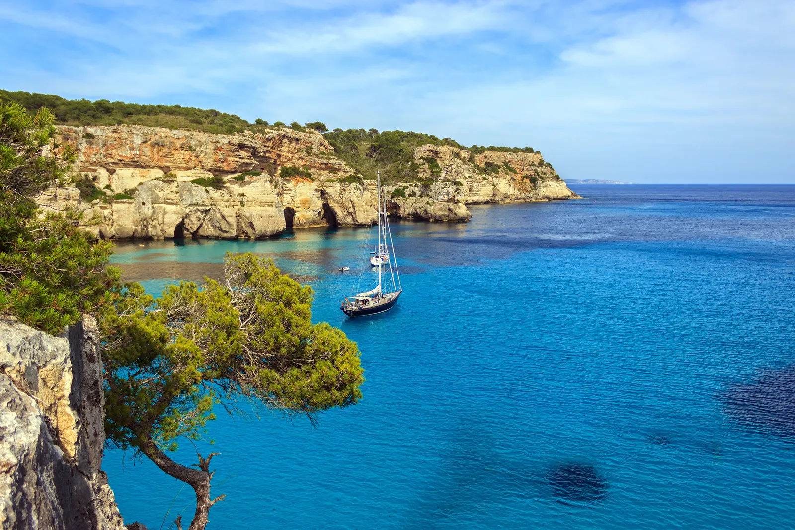 Velero fondeado en una cala de aguas turquesa bajo acantilados dorados en Menorca.